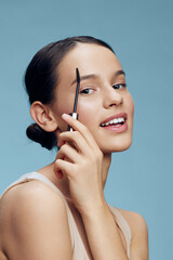 Fototapeta premium Young woman applying eyebrow makeup with a smile against a soft blue background, showcasing beauty and confidence in skincare routines