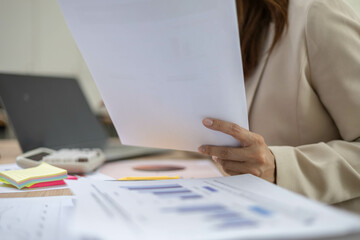 A woman is working at her desk with a laptop and a stack of documents. She is talking on the phone, looking at documents and seems focused on her work.

