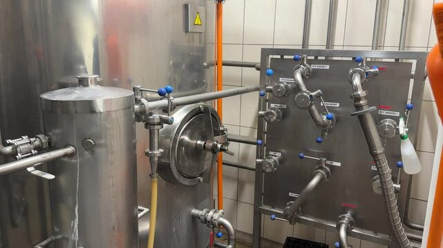 Stainless steel tank and labeled valve panel in a brewery, showing a network of pipes and connectors for managing liquid flow and processing stages.
