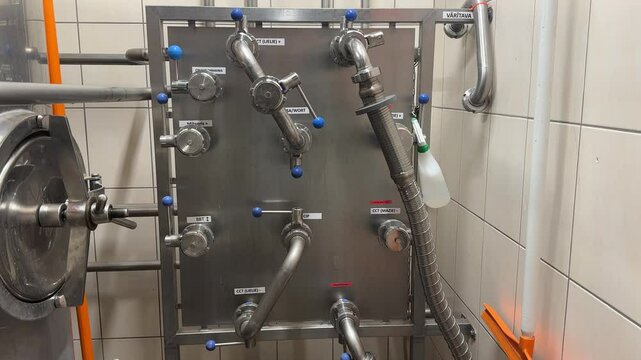 Stainless steel brewery valve panel with labeled pipe connections and flexible hose, used for controlling and directing liquid flow in production.
