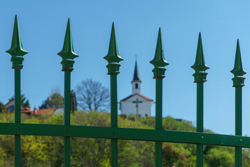 Fototapeta premium A small chapel on a distant mountain can be seen among the wrought iron fence pinnacles