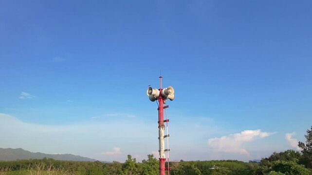 Outdoor warning siren pole with multiple loudspeakers against clear blue sky and mountain backdrop. Emergency disaster alert system for rural area safety, news broadcasting pole