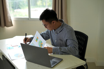 Young asian male officer is depressed while working on laptop and paper works on graphs, and charts and suffering from stress at office, he has to finish the data analysis in time before the deadline.