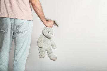 A person stands with their back to the viewer, casually holding a soft gray bunny toy by one ear in...