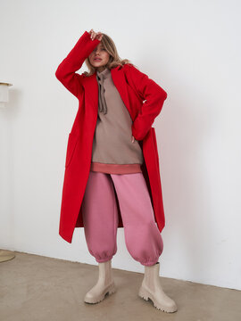 Fashionable woman poses in vibrant red coat covering her eyes with one arm; layered beige hoodie, pink baggy pants tucked into cream boots, standing on concrete floor in minimalist room