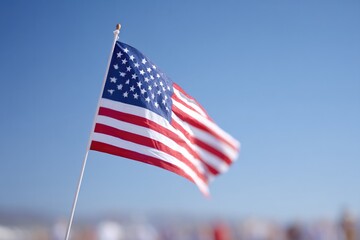 lone american flag waving against clear blue sky with blurred figures of people celebrating independence day in background