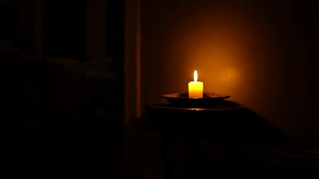 Illuminated short wax candle standing on dark table in dimly lit interior with warm glow, flame flickering against brown wall.