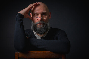 Thoughtful mature man with glasses and beard posing in studio with dramatic lighting. Conceptual portrait of wisdom, confidence, and introspection on black background