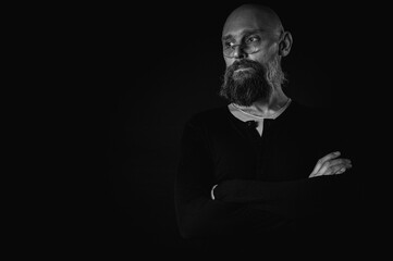 Black and white portrait, emotional bearded man in black hat and glasses, thoughtful look into the camera. Dramatic portrait in subdued lighting, expressive face, emotional tension, dark background.