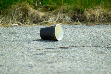 A black disposable cup lies on a gray asphalt surface close to dry grass. The scene captures the impact of littering in a natural environment during daylight