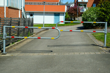 A metal barrier with a unique twisted design blocks access to a residential road. The tranquil neighborhood features vibrant greenery and sunny weather, highlighting the unusual obstacle
