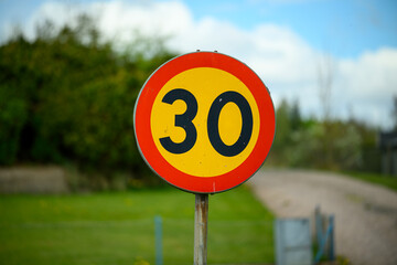 A large circular speed limit sign displays the number 30 in black on a red and yellow background. It stands prominently beside a well-maintained rural road under a blue sky