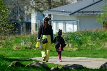 A mother and her young daughter stroll hand in hand along a paved path in a lush park. Bright sunshine illuminates their joyful outing amidst blooming nature