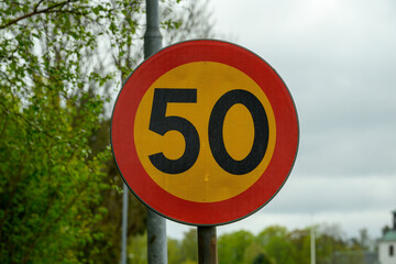 A circular speed limit sign displays the number 50 in bold black on a bright yellow background. The sign sits alongside a road surrounded by lush green trees, indicating a speed regulation area