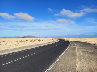Panoramic view of long empty desert asphalt road crossing El Corralejo Dunes Natural Park in Fuerteventura. Canary Islands. Lonely road in a desert area and blue sky. Driving and Landscape concept.