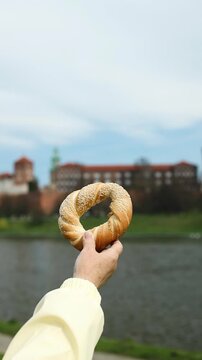 Hand holding a traditional Krakow bagel (obwarzanek) against the backdrop of the historic Wawel Castle and Vistula River. A perfect combination of local food, culture, and travel in Poland.
