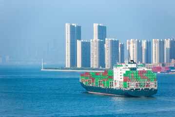 Fully loaded container ship departing Qingdao port, China. In the background, newly built waterfront residential towers rise along the shoreline.