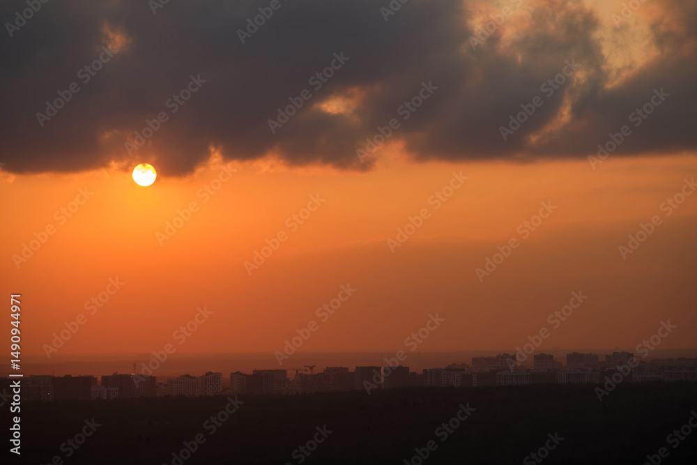 Wall mural sunset over city skyline with dark clouds. urban landscape with orange sky and setting sun. cityscap