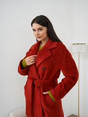 Plakat Woman with long dark hair wearing a bright red coat, fastened with a belt, over a green sweater. She stands with hand near collar, beside a tall lamp with a fringed shade