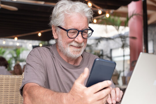 A smiling bearded senior man sits in a cafe using his laptop and smartphone. An elderly business man uses technology expressing confidence and contentment in a relaxed atmosphere.