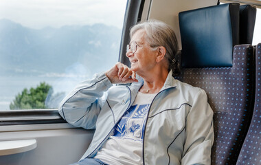 Senior smiling woman with eyeglasses enjoys the train ride sitting in passenger seat by the window looking out the landscape. Happy grandmother enjoying travel © luciano