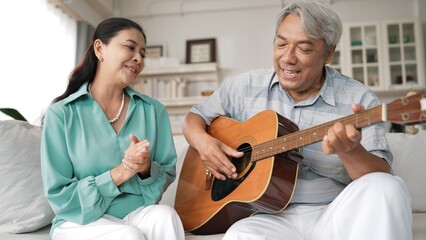 Obraz premium Happy senior man sitting at sofa and playing guitar or music instrument while his wife singing and moving to music. Elder couple spending time together and enjoy doing activity together. Myrmidon.