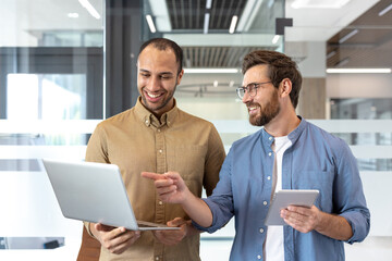 Two cheerful colleagues collaborate on a laptop, discussing ideas with the help of a tablet in a...