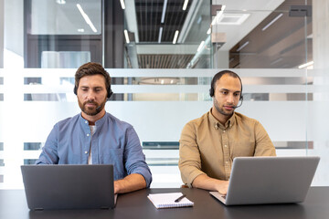Two business professionals wearing headsets work diligently on their laptops in a modern office setting. They focus on their tasks intently.