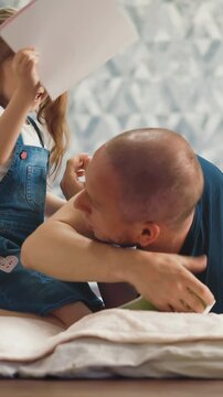 playful little girl with ponytails hits shaved bald father with notebook lying on floor with soft blanket in light room at quarantine time