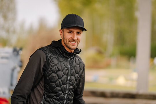 Man in a black cap and quilted vest smiling outdoors on a blurry park background during autumn or spring, with natural soft lighting.. - Powered by Adobe