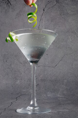 a dirty martini in a stemmed glass, garnished with a lime twist on the rim and a hand holding...
