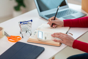 Asian businesswoman in red shirt is checking product orders and calculating sufficient stock to deliver to customers according to orders entered into online shopping application.Copy Space for text