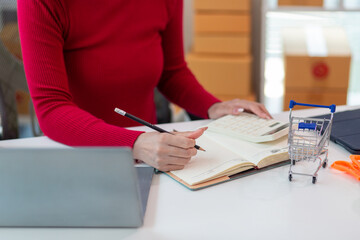 Asian businesswoman in red shirt is checking product orders and calculating sufficient stock to deliver to customers according to orders entered into online shopping application.Copy Space for text