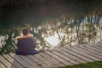 A child in a winter coat sits alone on a wooden dock, looking at the calm water reflecting trees and sunlight, creating a peaceful and contemplative mood