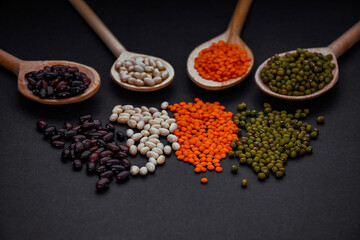 Variety of legumes in wooden spoons and scattered on black background. Healthy plant-based food ingredients. High-protein vegan products.