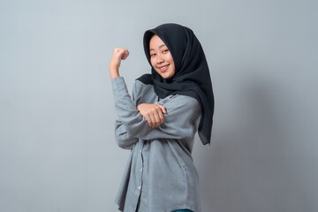 Confident young Asian Muslim woman in a black hijab and gray shirt showing biceps and smiling,...