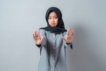 Serious young Asian Muslim woman wearing a hijab and gray shirt making a stop gesture with both...