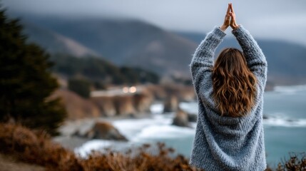 Obraz premium woman stands with her hands raised in a gesture of gratitude, overlooking a dramatic coastline. waves crash below, and mist hangs over the distant mountains, creating a serene atmosphere