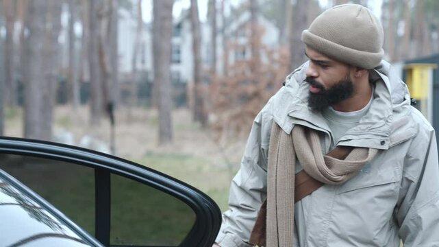 A handsome and smiling Black man in his early 30s with a thick beard in a light jacket approaches a black car, opens the door, and pulls out a bag of groceries on a terrace in the forest.