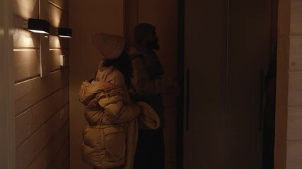 A woman walks and takes off her outerwear while a man walks next to her, opening the closet door and taking out two hangers