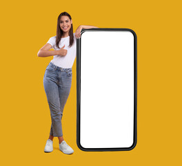 Cool Mobile Offer. Happy casual lady leaning on huge cellphone with empty white screen, showing thumb up, recommending great new app or website for cell phone, offering space for your ad, mock up