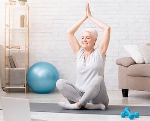 Online training. Calm senior woman practicing yoga at home with laptop, meditating and doing...