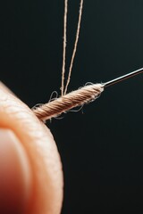 Close up view of thread being pulled through eye of needle, detail of beige yarn and metal with dark background.