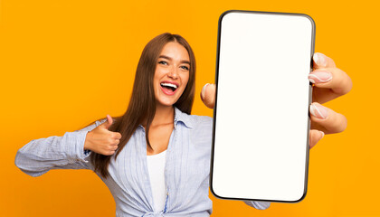 Recommendation. Portrait of excited lady holding big smartphone with white blank screen in hand, showing close up to camera making thumb up gesture. Gadget with empty free space for mock up, banner
