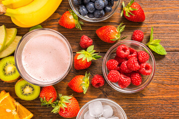 Top view of glass with summer strawberry smoothie surrounded by fresh berries bananas kiwi orange and mint leaves on rustic wooden table with copy space
