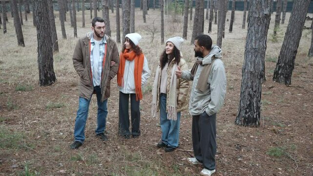 A group of four friends of different ethnicities in their 30s standing in the forest surrounded by a few trees, looking around and talking, two women are in beanies and jackets while two men are in ja