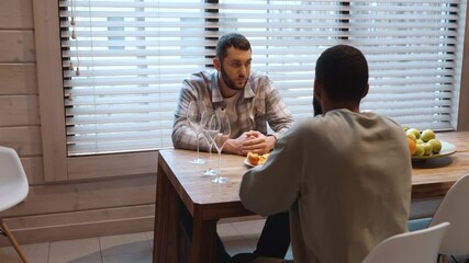 A Caucasian man in his 30s with short dark hair is talking with an adult Black man while they are sitting at a wooden table with wine glasses and a plate of a tangerine in a room with a big window