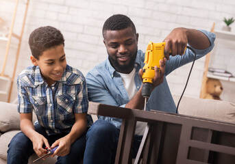 Black father teaching his son how to use drill, perforating chair in living room at home, sharing...