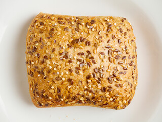 One crusty roll with seeds and grain on white plate on simple light color table cloth. Fine bakery product made from high quality ingredients, Small bread.