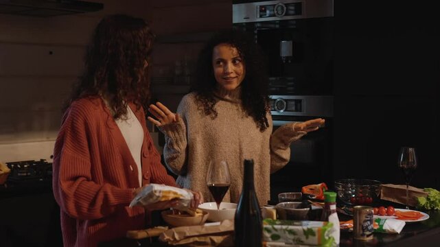 A woman is talking to a female friend who is pouring breadsticks into a bowl, they are laughing while standing at the table